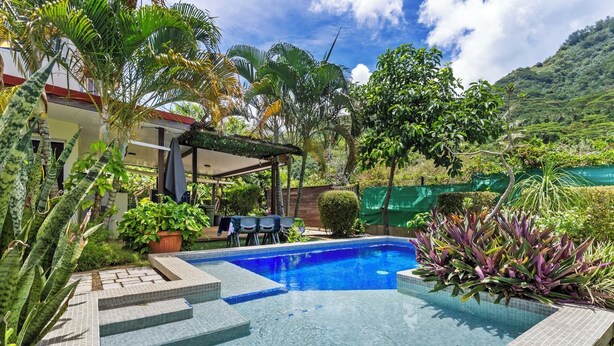 Una piscina con escalones, rodeada de plantas tropicales y un porche cubierto.