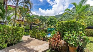Un jardín con un sendero de hormigón, diversas plantas, una piscina pequeña y un paisaje montañoso de fondo.