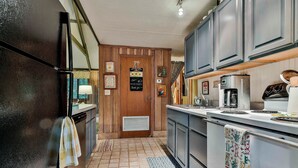 Una cocina con refrigerador, estufa y alacenas. Se observa una puerta con un letrero, una ventana y una alfombra estampada.