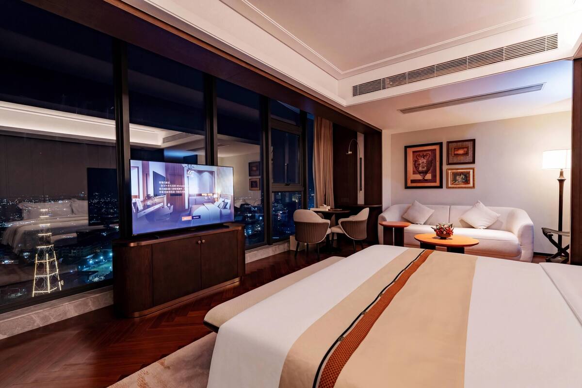 A hotel room with a large bed, a TV, a sofa, a desk, and a view of the city at night.