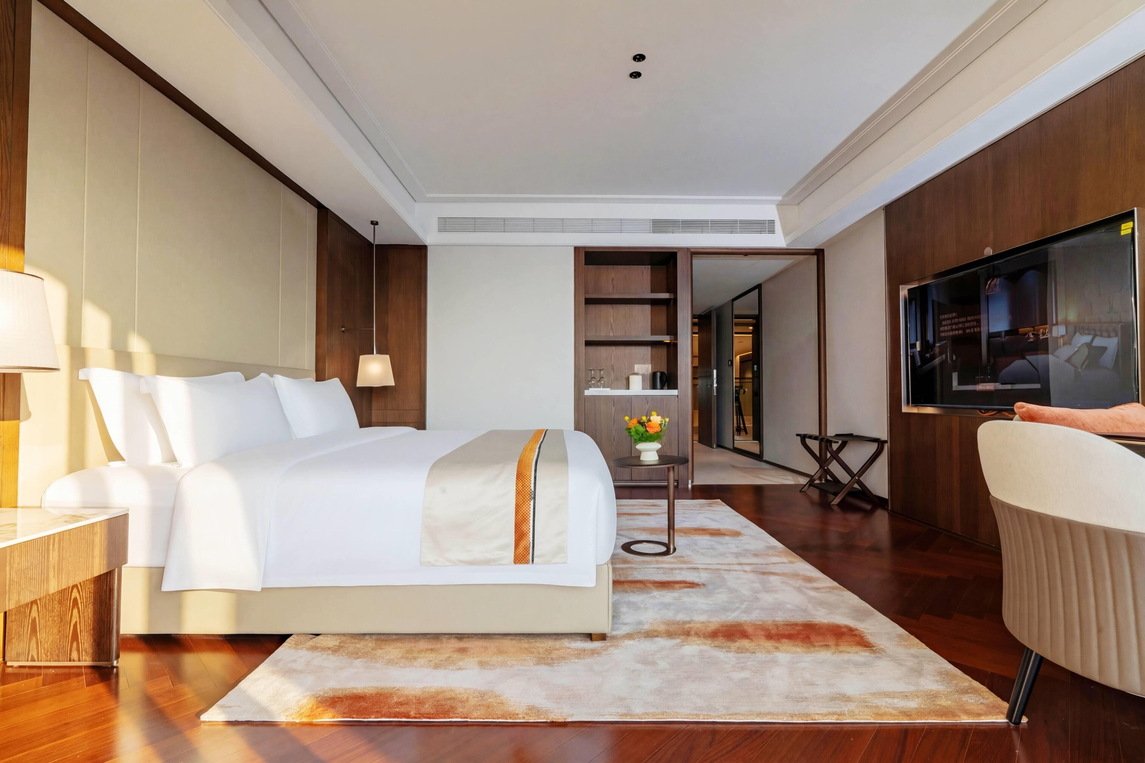 Ein modernes Hotelzimmer mit einem großen Bett, einem Fernseher, einem Nachttisch, einem kleinen Beistelltisch mit einer Vase und Holzboden.
