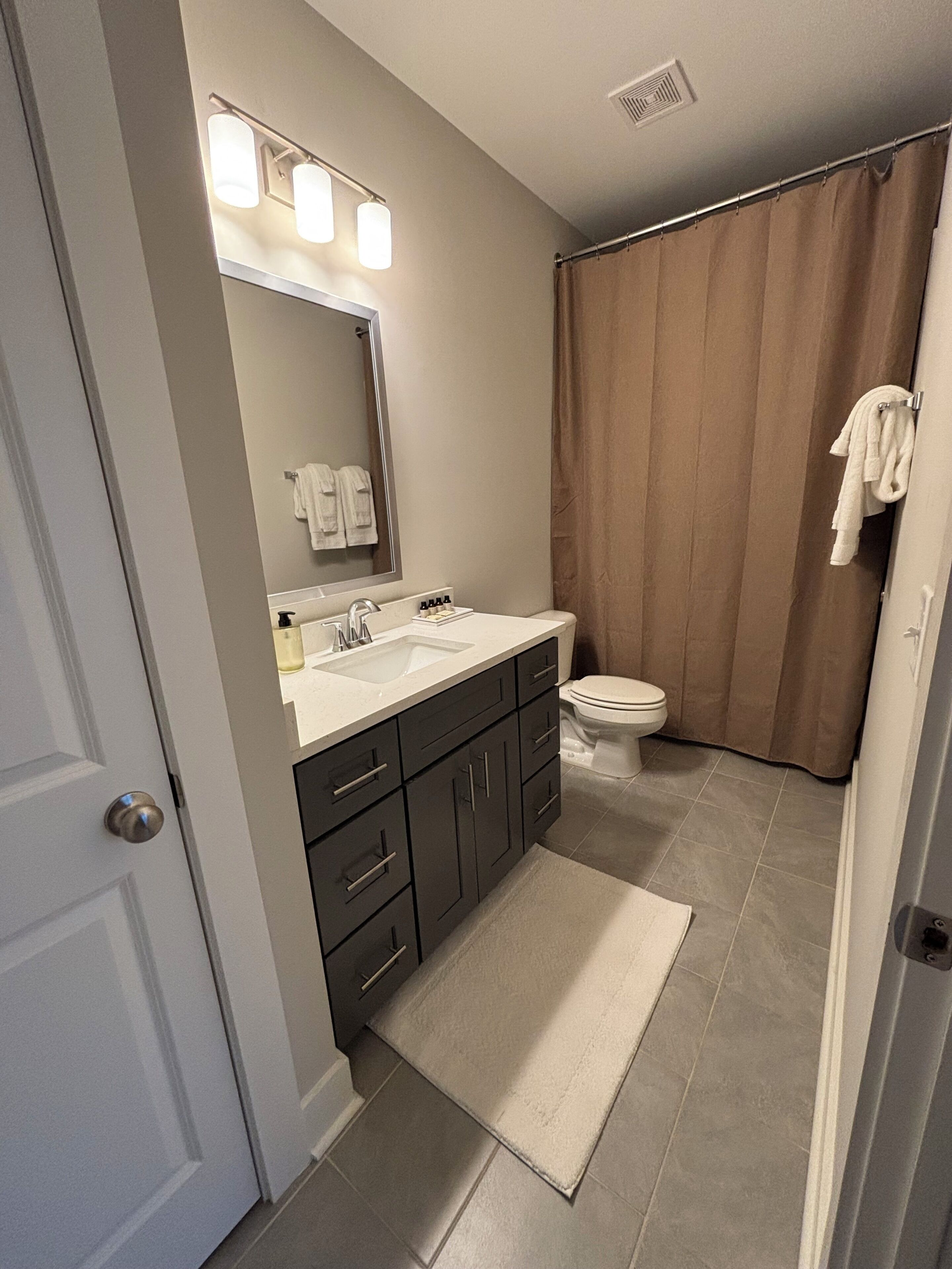 A modern bathroom with a dual sink vanity, a toilet, a shower curtain, and a large mirror with overhead lighting.