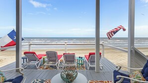 Un patio frente a la playa con sillas blancas y cojines rojos, una mesa con un centro de mesa decorativo y vista al océano.