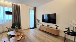 Moderni olohuone, jossa on puulattia, musta tuoli, puinen ruokapöytä, seinälle asennettu televisio ja ruukussa oleva kasvi.