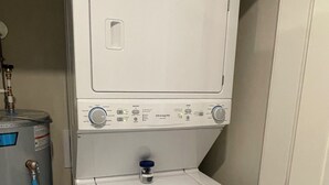 Un appareil compact, blanc, comprenant une laveuse et une sécheuse, conçu pour être fixé au mur, avec un panneau de commande, un réservoir d’eau et un tuyau.
