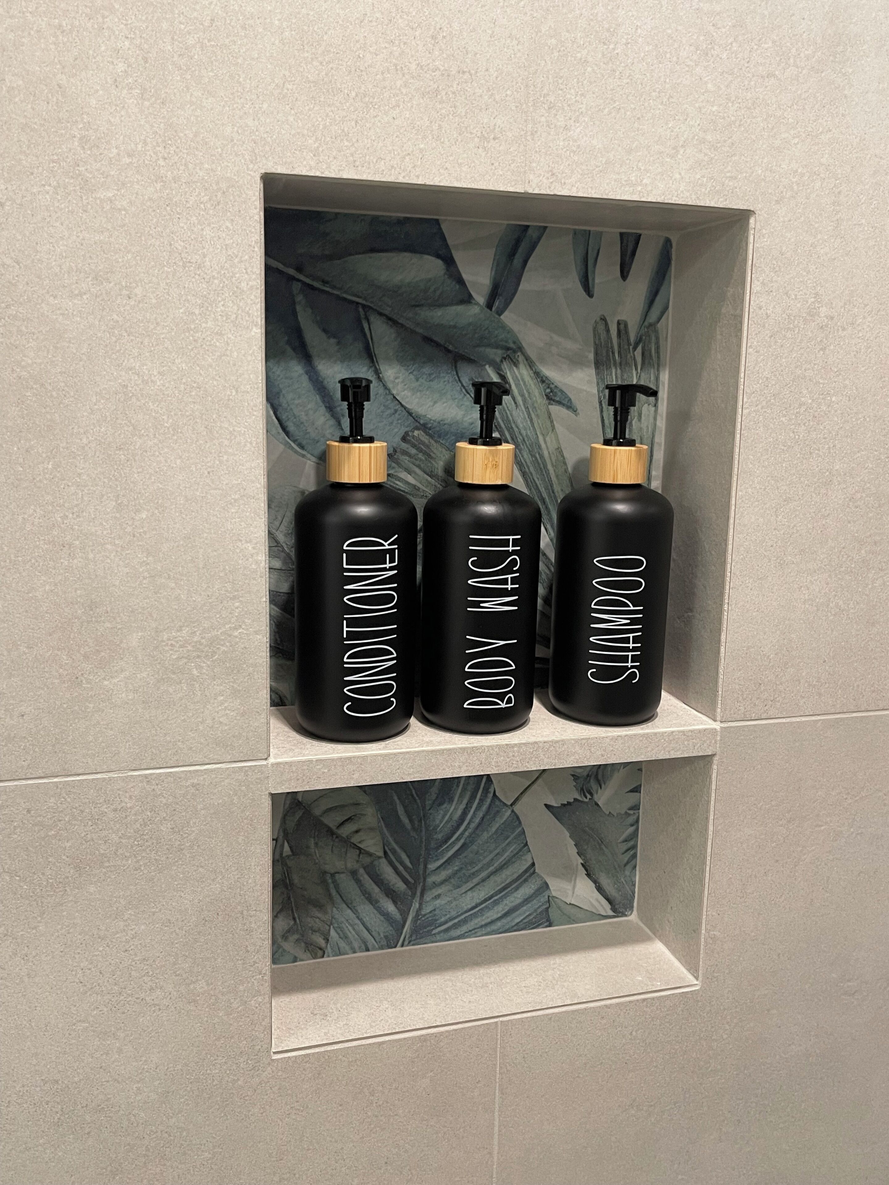 Drei schwarze Flaschen mit Holzverschlüssen und Pumpspendern, beschriftet mit 'CONDITIONER', 'BODY WASH' und 'SHAMPOO', auf einem Flaschenregal in einem gefliesten Badezimmer.