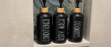 Three black bottles with wooden caps and pump dispensers, labeled 'CONDITIONER,' 'BODY WASH,' and 'SHAMPOO,' in a tiled bathroom shelf.