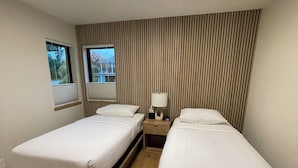 Une chambre d’hôtel avec deux lits simples, une table de chevet avec une lampe, une fenêtre avec des stores et un mur à rayures.