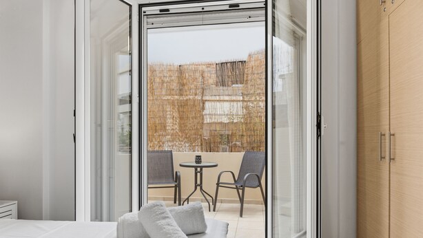 A modern hotel room with a bed, a small table, two chairs, and a sliding glass door leading to an outdoor area with a bamboo fence.
