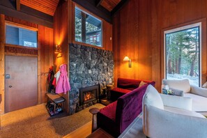 Ein gemütliches Zimmer mit Kamin aus Stein, Holzwänden und einem großen Fenster mit Blick auf eine verschneite Landschaft.