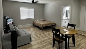 Ein Schlafzimmer mit einem Bett, einem Deckenventilator, einem Fenster mit Jalousien, einem Nachttisch, einem Fernseher, einem grauen Sofa und einem Holzessttisch mit Stühlen.