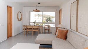 Een moderne woonkamer met een bank, eettafel en stoelen, een groot raam met uitzicht, en een hanglamp.
