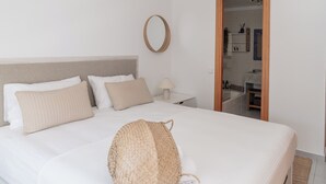 Een bed met witte lakens, een geweven tas en een nachtkastje met een lamp.