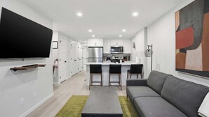 Una cocina moderna con armarios blancos, un refrigerador de acero inoxidable y un microondas. Hay un comedor con un sofá gris y una mesa pequeña.
