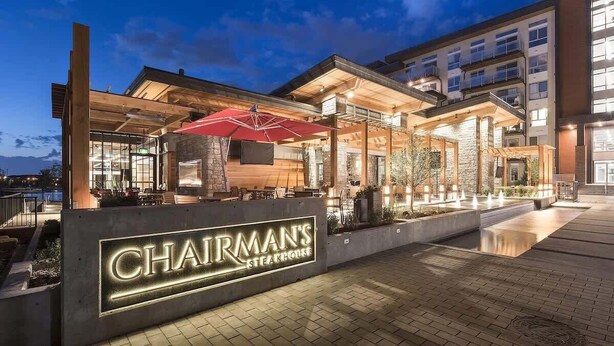 Un edificio moderno con un letrero que dice 'Chairman's Steakhouse', mesas al aire libre y fachada de piedra.