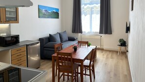 A compact kitchen with a microwave, refrigerator, and wooden cabinets. A gray sofa and dining table with chairs are positioned near a window with curtains. - Prague Deluxe City Center Apartment (Hlavní město Praha)