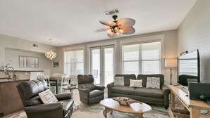 Un salon moderne comprenant un canapé, un fauteuil et une table basse, un coin repas avec des chaises, une cuisine avec un évier et un ventilateur de plafond.