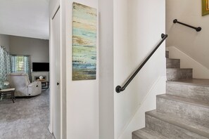 Una escalera con barandillas de metal, una sala de estar con un sofá color beige y un televisor de pantalla plana, y un cuadro en la pared.
