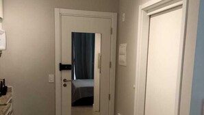 Un couloir étroit avec une porte donnant sur une pièce, une petite armoire contenant des objets sur le côté gauche, et un interrupteur sur le mur.