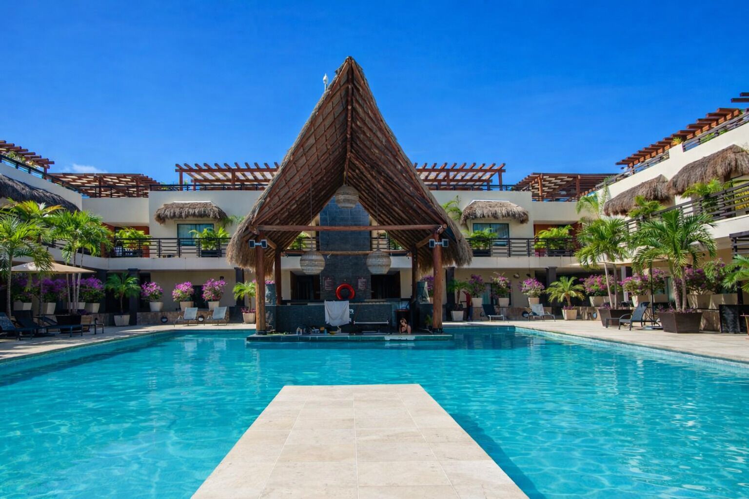 A swimming pool with a thatched-roof structure in the center, surrounded by lounge chairs and potted plants.