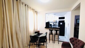 A dining area with a table and chairs, a kitchen in the background, and a living area with a sofa and a TV. - Modern Apartment in the South Zone (Uberlândia)