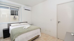 A bedroom with a bed, a window, an air conditioning unit, a door, and a bedside table. - Apartamento na Trindade - CEN008 (Florianópolis)