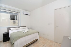 A bedroom with a bed, a window, an air conditioning unit, a door, and a bedside table. - Apartamento na Trindade - CEN008 (Florianópolis)