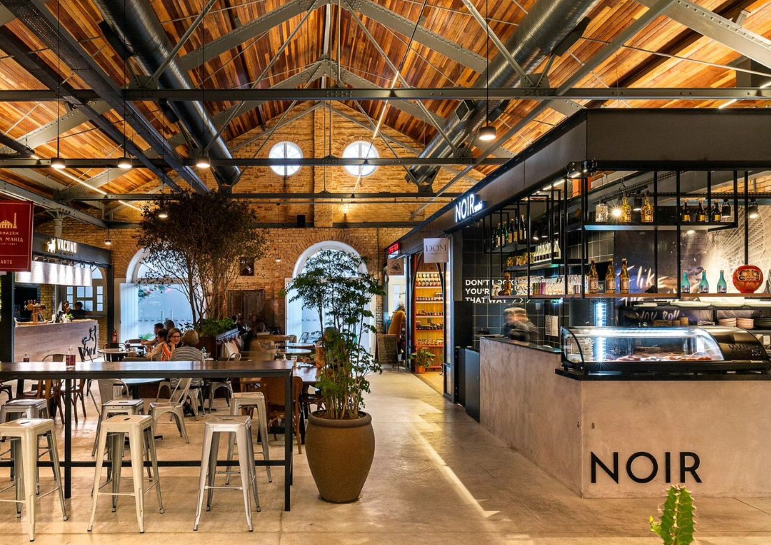 Interior de un restaurante con techo alto, vigas a la vista y una zona de bar identificada como 'NOIR'.