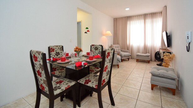 A dining area with a glass table, floral chairs, and a living area with a sofa and curtains. - Apartamento na Trindade - CEN008 (Florianópolis)