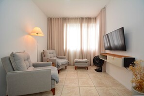 A living room with a sofa, armchair, ottoman, floor lamp, TV, and a fan. - Apartamento na Trindade - CEN008 (Florianópolis)