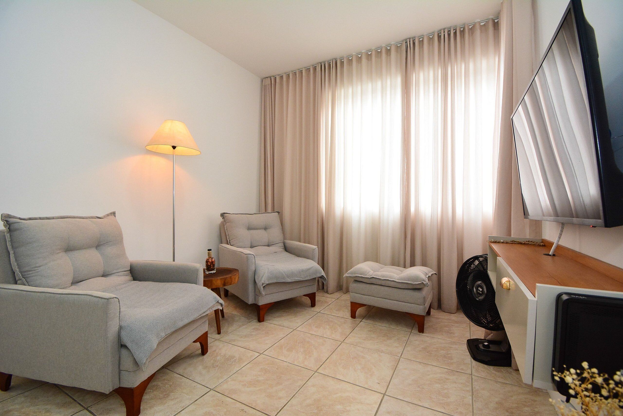 A living room with a sofa, armchair, and ottoman, a standing lamp, a flat-screen TV, and a fan.