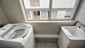 A small bathroom with a washing machine, a sink, and a window with a view of buildings. - Apartamento na Trindade - CEN008 (Florianópolis)
