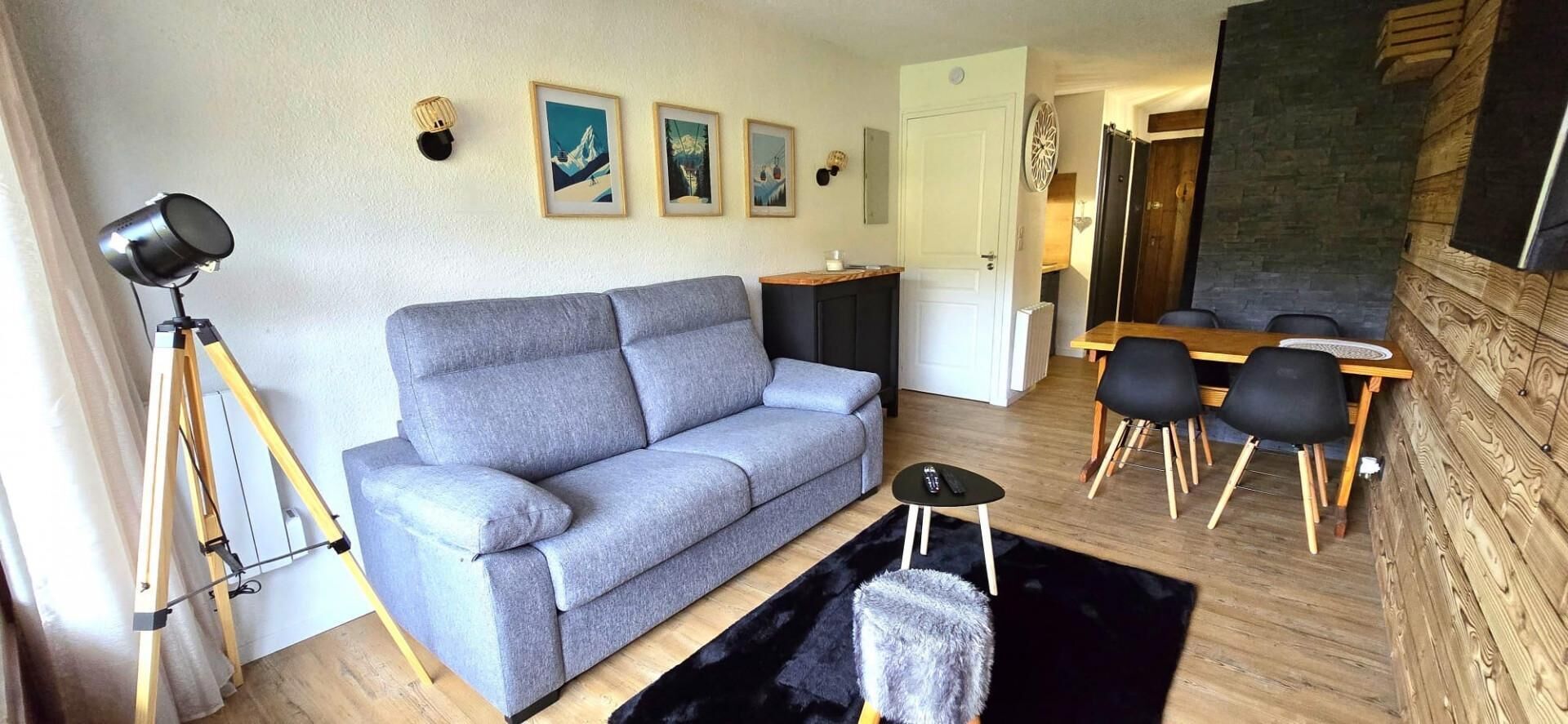 Ein Wohnzimmer mit grauem Sofa, einem kleinen schwarzen Hocker, einem Stativ mit einer Kamera und einem Essbereich mit Holztisch und schwarzen Stühlen.