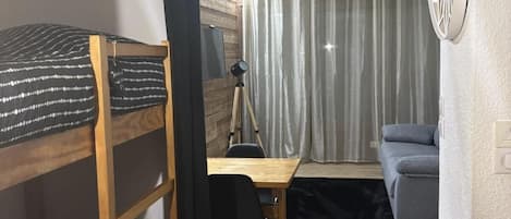 Ein kleines Zimmer mit Etagenbetten, einem Schreibtisch, einem Stuhl, einem Heizkörper und einem grauen Sofa.