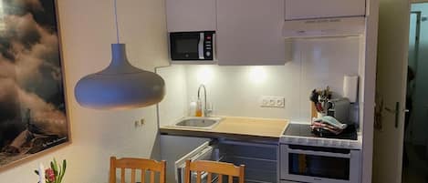 Cocina compacta con microondas, horno y lavaplatos. Hay una mesa de comedor con sillas y una lámpara colgante encima.