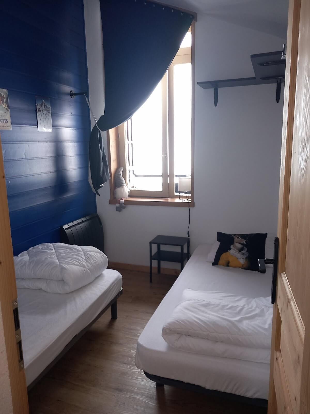 Una habitación con dos camas, una ventana con cortinas, una mesita pequeña y un estante fijado a la pared.