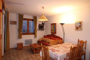 Ein gemütliches Zimmer mit Bett, Tisch, Stühlen, einem Fenster mit Vorhängen, einer Heizung und einer Lampe.
