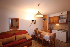 Ein kompakter Wohnraum mit Sofa, Esstisch, Stühlen, einer Küchenzeile inklusive Mikrowelle und Herd sowie einer Pendelleuchte.