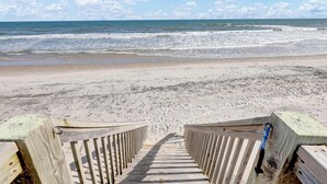 Ein hölzerner Steg führt zu einem Strand mit Wellen und blauem Himmel.