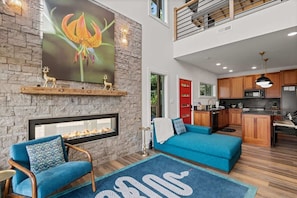 A modern living room with a fireplace, a blue sofa, and a kitchen area. - Heavenly Views Hot Tub Games Killer Design (Sevierville)