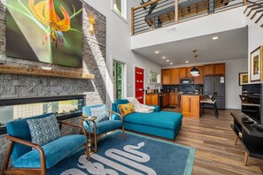 A modern living space with a stone fireplace, a kitchen with wooden cabinets, and a dining area with a blue sofa. - Sunset Mountain Views · Hot Tub · Gated (Sevierville)