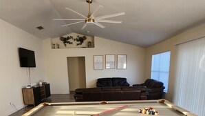 Une table de billard dans une pièce dotée d’un ventilateur de plafond, d’un téléviseur, d’un canapé et d’œuvres d’art encadrées accrochées au mur.