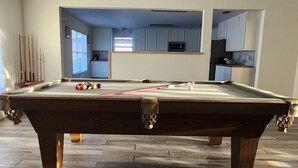Une table de billard dans une pièce dont le plancher est en bois avec un motif en chevrons, un coin cuisine à l’arrière-plan et un plafond doté d’un éclairage encastré.
