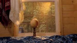 A wooden cabin room with a large window, a bed with a patterned bedspread, and a wall-mounted lamp.