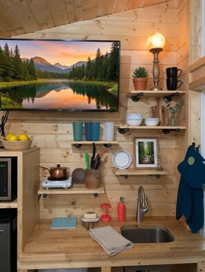 A wooden cabin interior with a television, shelves with kitchenware, a sink, and a window overlooking a lake.