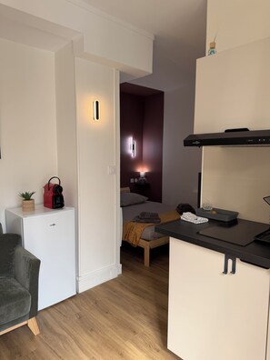 Ein kompakter Wohnraum mit Küchenzeile, einem Bett mit Kopfteil, einer kleinen Sitzecke und einer Tür zu einem anderen Raum.