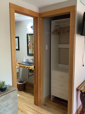 A room with a wooden floor, a white closet with hanging space, a small sink with a mirror, and a wooden cabinet.
