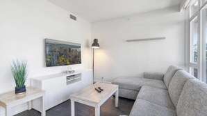 Una sala moderna con un sofá gris, una pequeña mesa de centro blanca, una lámpara de pie, un cuadro en la pared y una planta en maceta.