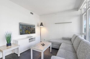 Ein modernes Wohnzimmer mit einem grauen Sofa, einem kleinen weißen Couchtisch, einer Stehlampe, einem Bild an der Wand und einer Topfpflanze.