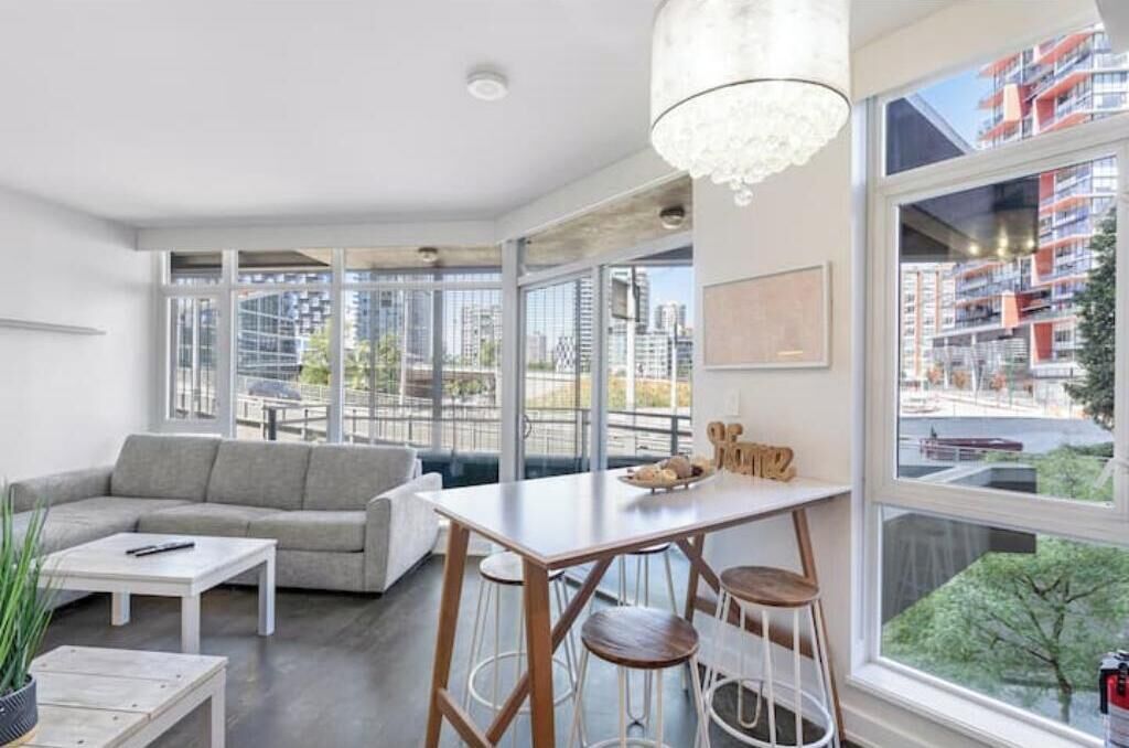 A modern living room with a sofa, coffee table, and bar stools. Large windows offer a view of the cityscape.